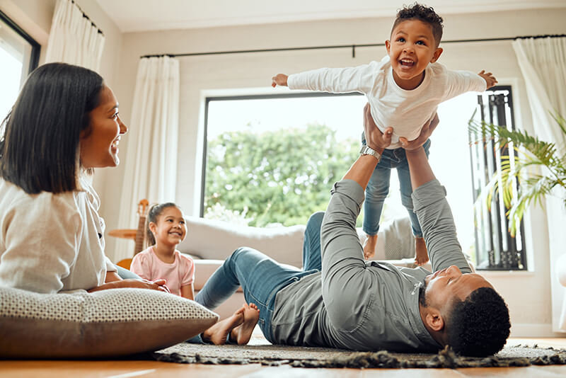 A joyful family plays together in a bright living room, with a child being lifted in the air by a parent while others watch and smile.